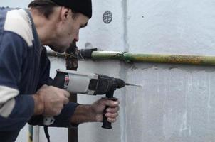 un anziano operaio trapani un' buco nel un' polistirolo parete per il successivo installazione di un' plastica rafforzando tassello. la creazione di fori nel il parete con un' trapano. riscaldamento di il edificio facciata foto