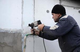 un anziano operaio trapani un' buco nel un' polistirolo parete per il successivo installazione di un' plastica rafforzando tassello. la creazione di fori nel il parete con un' trapano. riscaldamento di il edificio facciata foto