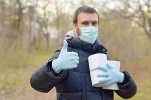 covidiot concetto. giovane uomo nel protettivo maschera detiene molti rotoli di gabinetto carta e Spettacoli pollice su all'aperto nel primavera legna foto