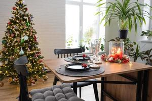 festivo tavolo impostato nel il vivente camera per Natale e nuovo anno nel soffitta stile. Natale albero, nero piatti e forchette, intrecciata tovaglioli, di moda stoviglie, accogliente interno di il Casa foto