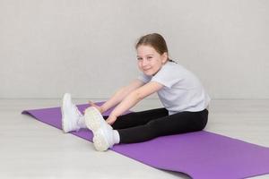 poco carino ragazza si siede su un' ginnastica stuoia e fa allungamento esercizi, tira sua mani per sua piedi foto