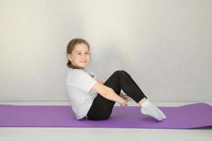 carino bambino si siede su un' ginnastica stuoia con sua ginocchia piegato e sua piedi teso fuori, il bambino sorrisi foto