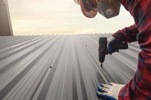 vicino su trapano roofer lavoratore nel protettivo uniforme indossare e guanti, coperture strumenti, installazione nuovo tetti sotto costruzione, elettrico trapano Usato su nuovo tetti con metallo foglio. foto