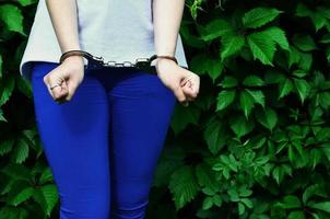 frammento di un' giovane penale ragazze corpo con mani nel manette contro un' verde fioritura edera le foglie sfondo. il concetto di trattenendo un colpevole di un' femmina penale nel un' rurale ambiente foto