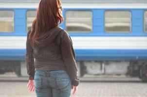 un' giovane dai capelli rossi ragazza è in piedi su il ferrovia piattaforma e Guardando il in partenza treno. il donna era in ritardo per sua treno. indietro Visualizza foto