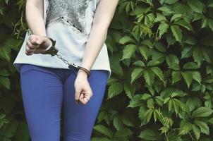 frammento di un' giovane penale ragazze corpo con mani nel manette contro un' verde fioritura edera le foglie sfondo. il concetto di trattenendo un colpevole di un' femmina penale nel un' rurale ambiente foto