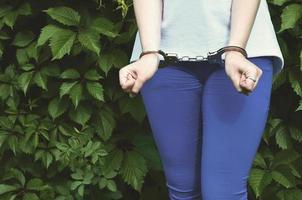 frammento di un' giovane penale ragazze corpo con mani nel manette contro un' verde fioritura edera le foglie sfondo. il concetto di trattenendo un colpevole di un' femmina penale nel un' rurale ambiente foto