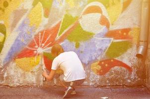 foto di un' giovane tipo nel denim pantaloncini e un' bianca camicia. il tipo disegna su il graffiti parete un' disegno con aerosol vernici di vario colori. il concetto di teppismo e danno per proprietà