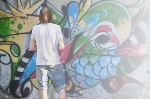 foto di un' giovane tipo nel denim pantaloncini e un' bianca camicia. il tipo disegna su il graffiti parete un' disegno con aerosol vernici di vario colori. il concetto di teppismo e danno per proprietà