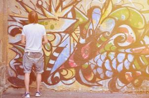 foto di un' giovane tipo nel denim pantaloncini e un' bianca camicia. il tipo disegna su il graffiti parete un' disegno con aerosol vernici di vario colori. il concetto di teppismo e danno per proprietà