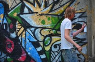 foto di un' giovane tipo nel denim pantaloncini e un' bianca camicia. il tipo disegna su il graffiti parete un' disegno con aerosol vernici di vario colori. il concetto di teppismo e danno per proprietà