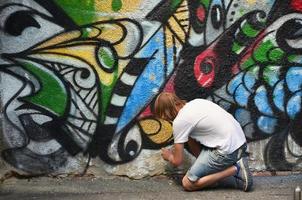 foto nel il processi di disegno un' graffiti modello su un vecchio calcestruzzo parete. giovane dai capelli lunghi biondo tipo disegna un astratto disegno di diverso colori. strada arte e vandalismo concetto