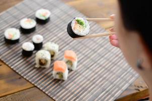 brunetta ragazza con bastoncini detiene un' Sushi rotolo su un' bambù cannuccia servire stuoia sfondo. tradizionale asiatico cibo foto