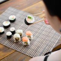 brunetta ragazza con bastoncini detiene un' Sushi rotolo su un' bambù cannuccia servire stuoia sfondo. tradizionale asiatico cibo foto