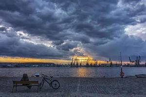 un' uomo con un' bicicletta seduta su un' panchina nel un' porto per ammirare il tramonto o Alba foto