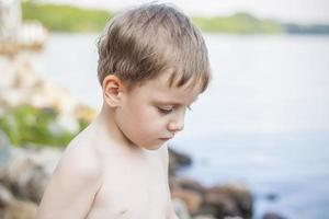 un' carino biondo ragazzo esperienze il gioia di a piedi su un' estate giorno di il fiume, seduta su il succoso erba. divertente facciale espressione. estate calore, un' bambino senza un' maglietta. foto