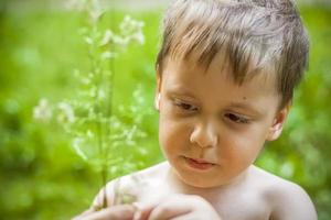 un' carino biondo ragazzo esperienze il gioia di a piedi su un' estate giorno di il fiume, seduta su il succoso erba. divertente facciale espressione. estate calore, un' bambino senza un' maglietta. foto