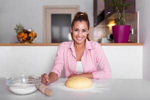 bellissimo giovane donna guardare a telecamera e sorridente mentre cottura al forno nel cucina a casa. sorridente giovane casalinga fabbricazione Impasto foto