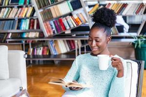 africano americano ragazza lettura un' libro sorridente contento e rilassato su un' tempo libero giorno a casa. casa biblioteca con libreria nel il Indietro. foto