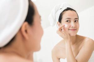 contento giovane asiatico donna l'applicazione viso lozioni mentre indossare un' asciugamano e toccante sua viso nel bagno foto