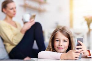 giovane mamma e sua poco figlia utilizzando smartphone e seduta su divano a casa foto