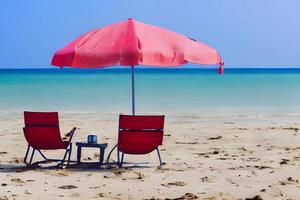 un' coppia di rosso sedie seduta sotto un' rosa ombrello su il spiaggia. generativo ai. foto