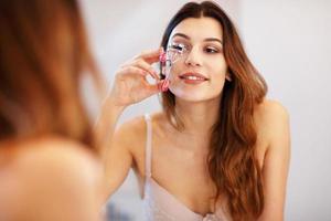 attraente giovane donna fare trucco mentre guardare a il specchio nel bagno foto