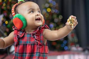 africano americano bambino è felicemente sorridente come giocando con ornamento fronzolo mentre condimento nel Natale vestito e Santa cappello con Natale albero su il indietro per stagione celebrazione concetto foto