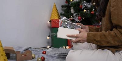 sorridente asiatico donna nel festivo umore Tenere avvolto regalo scatola con Marrone nastro nel mani, allungamento su presente per telecamera, festeggiare allegro Natale miracolo tempo o nuovo anno a casa. foto