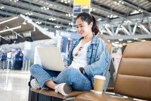 asiatico femmina viaggiatore nel casuale Abiti è utilizzando il computer portatile nel il partenza sala a il internazionale aeroporto su un' fine settimana viaggio. foto