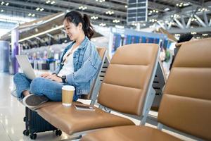asiatico femmina turisti casuale stoffa con zaino viaggiatore rilassare sedersi aspettare nel aeroporto terminale, lei uso il computer portatile. sicurezza viaggio concetto foto