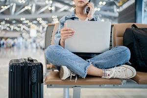 viaggio vacanza concetto, asiatico femmina donna usi un' smartphone e è Lavorando su un' computer portatile, lei sedersi wating per volo programma a terminale aeroporto. foto