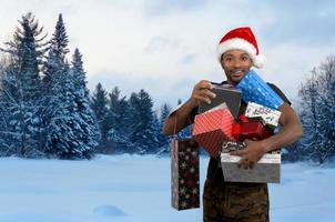 uomo Tenere regalo scatole per Natale nel il neve inverno paesaggio e indossare rosso Santa cappello al di fuori foto