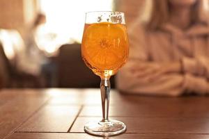 un' bicchiere di aperol e un' fetta di arancia su un' tavolo nel un' bar. nel il sfondo, un' sfocato figura di un' donna seduta a un' tavolo foto