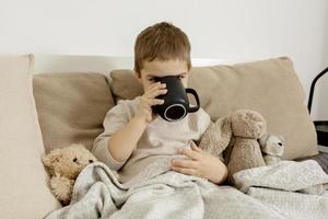 malato poco ragazzo potabile caldo tè su il letto a casa. male, malattia bambino avvolto nel un' lenzuolo, con boccale nel il suo camera. influenza stagione. interno e Abiti nel naturale terra colori. accogliente ambiente. foto