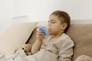 malato poco ragazzo con inalatore per tosse trattamento. indisposto ragazzo fare inalazione su il suo letto. influenza stagione. medico procedura a casa. interno e Abiti nel naturale terra colori. accogliente ambiente. foto