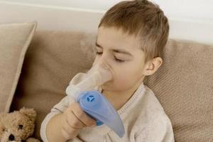 malato poco ragazzo con inalatore per tosse trattamento. indisposto ragazzo fare inalazione su il suo letto. influenza stagione. medico procedura a casa. interno e Abiti nel naturale terra colori. accogliente ambiente. foto