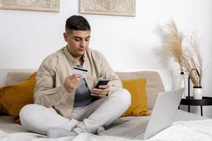 giovane uomo sorridente mentre fare surf il ragnatela su il suo smartphone e Tenere credito carta nel il suo mano provando per acquistare qualcosa in linea. facile in linea shopping a casa, commercio elettronico. accogliente interno. foto