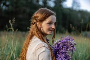 vicino su ritratto di giovane bellissimo testa Rossa donna con lentiggini, indossare bianca vestire, in posa nel il natura. ragazza con rosso capelli Tenere fiori. naturale bellezza. diversità, individuale unicità. foto