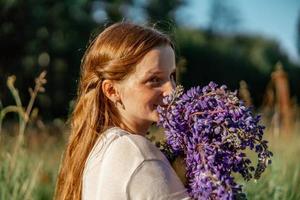 vicino su ritratto di giovane bellissimo testa Rossa donna con lentiggini, indossare bianca vestire, in posa nel il natura. ragazza con rosso capelli Tenere fiori. naturale bellezza. diversità, individuale unicità. foto