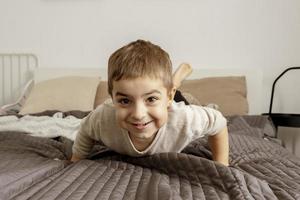 ritratto di piccolo, sorridente caucasico ragazzo su il letto a casa. carino bambino rilassante, riposo nel Camera da letto. positivo emozioni. accogliente e moderno interno. naturale, terra colori. casuale Abiti. foto