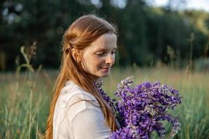 vicino su ritratto di giovane bellissimo testa Rossa donna con lentiggini, indossare bianca vestire, in posa nel il natura. ragazza con rosso capelli Tenere fiori. naturale bellezza. diversità, individuale unicità. foto