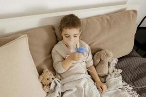 malato poco ragazzo con inalatore per tosse trattamento. indisposto ragazzo fare inalazione su il suo letto. influenza stagione. medico procedura a casa. interno e Abiti nel naturale terra colori. accogliente ambiente. foto