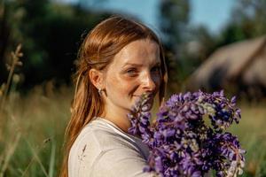 vicino su ritratto di giovane bellissimo testa Rossa donna con lentiggini, indossare bianca vestire, in posa nel il natura. ragazza con rosso capelli Tenere fiori. naturale bellezza. diversità, individuale unicità. foto