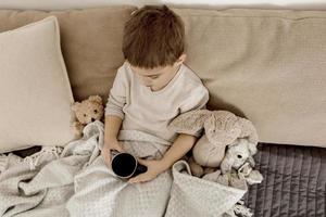 malato poco ragazzo potabile caldo tè su il letto a casa. male, malattia bambino avvolto nel un' lenzuolo, con boccale nel il suo camera. influenza stagione. interno e Abiti nel naturale terra colori. accogliente ambiente. foto