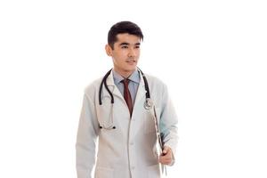 bello giovane brunetta uomo medico nel bianca uniforme con stetoscopio guardare a parte isolato nel studio foto
