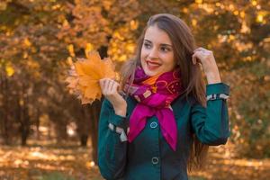 sorridente bella ragazza nel rosa sciarpa sta nel il parco si aggrappa per il capelli mentre il altro mano detiene il le foglie foto