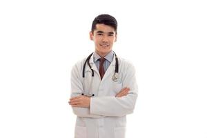 bella giovane brunetta uomo medico nel bianca uniforme con stetoscopio guardare e sorridente su telecamera isolato nel studio foto