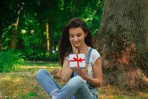 bellissimo giovane ragazza seduta su il erba e detiene un' piccolo regalo nel il mani foto