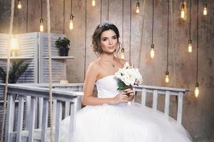 giovane contento sposa indossare bellissimo lussureggiante vestito nel il camera con un' lotto di leggero bulbi foto